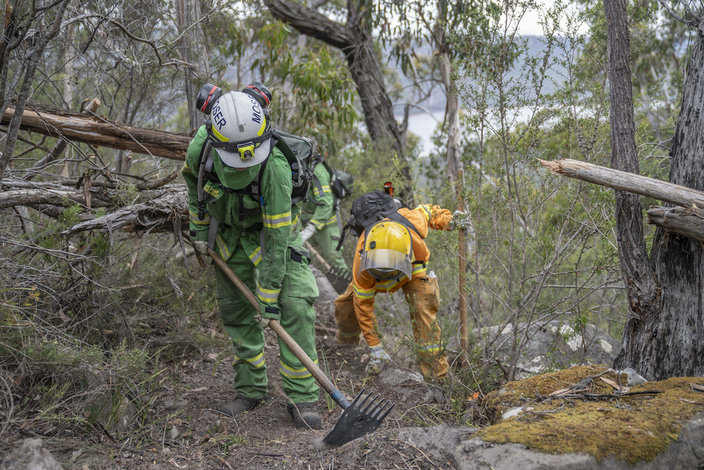 A firefighter in green fire clothes holds a rakehoe while standing on rocky terrain. In the background another fire fighter in orange bends down to pull some scrub away. 