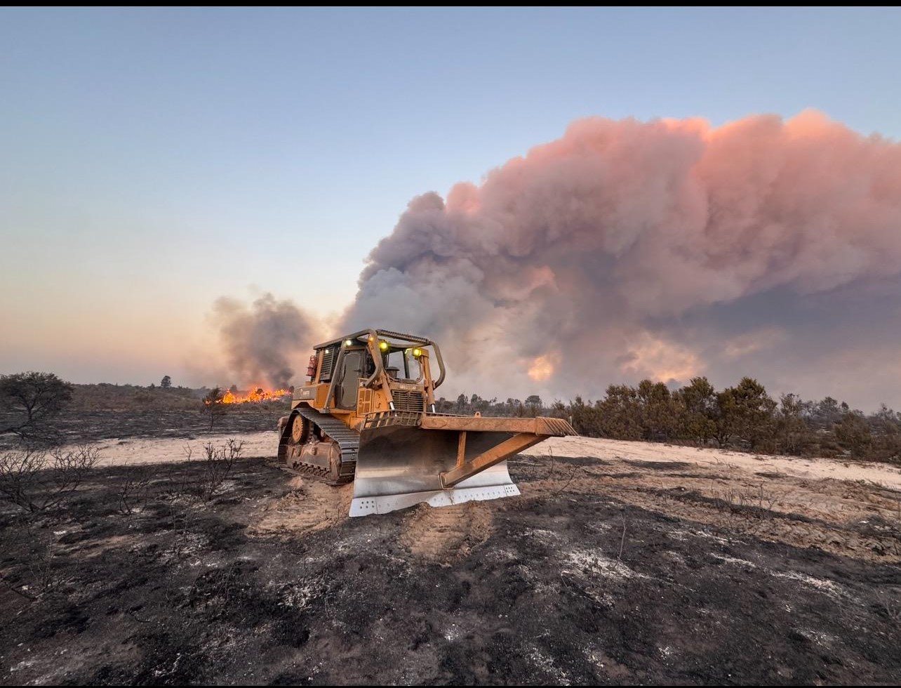 Photo of bulldozer in foreground with billowing purple-grey smoke in the background