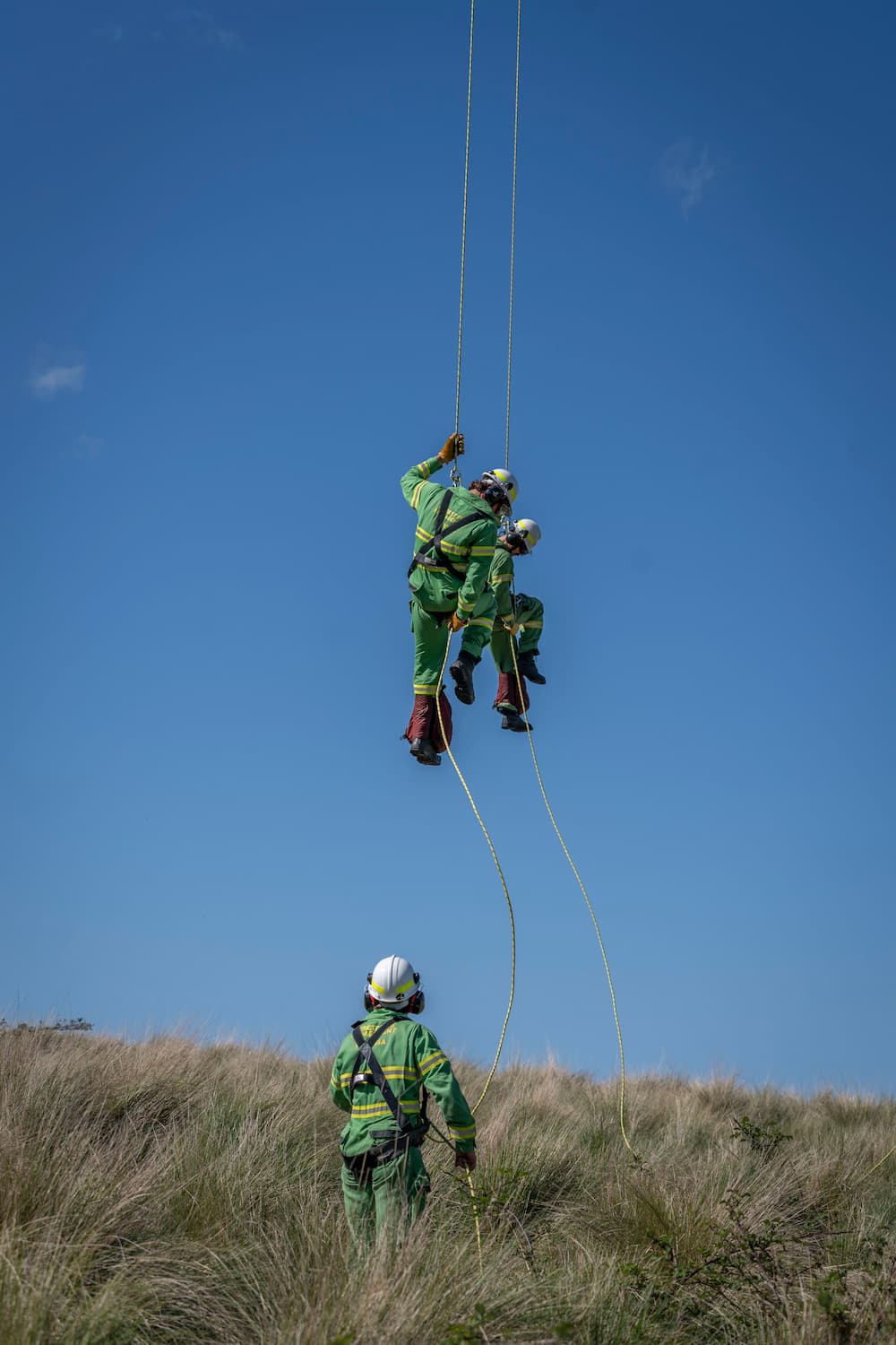 Two firefighters rappel to ground with clear blue skies in the background, one firefighter is already on the ground grasslands, wearing a helmet and looking up.