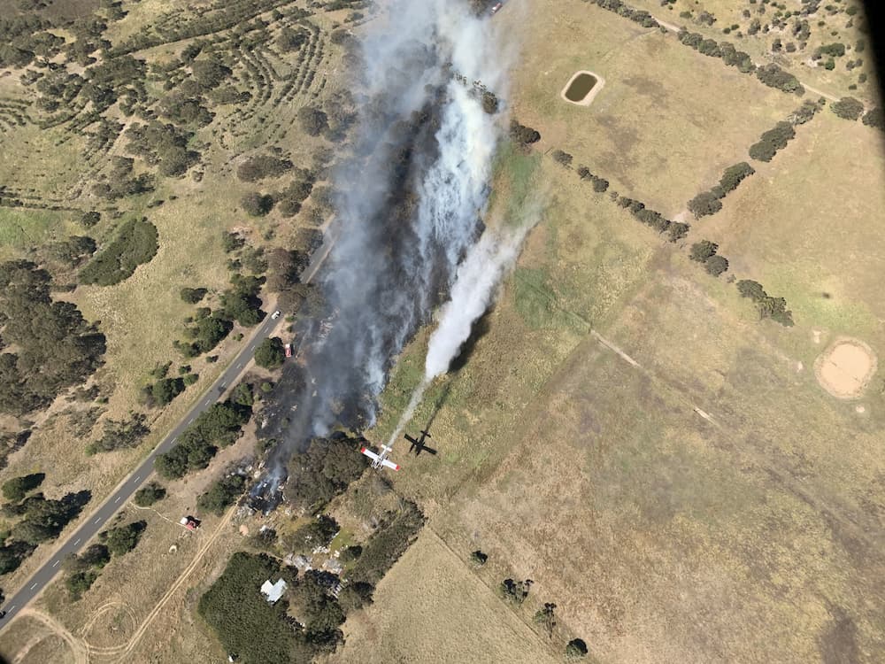 Bird's eye view of fixed-wing aircraft flying over a small fire in grassland near a road. Grey smoke is billowing from the ground