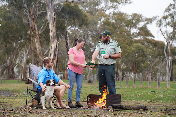 Man sits withdog by campfire, next to them a woman is standing speaking to a compliance officer